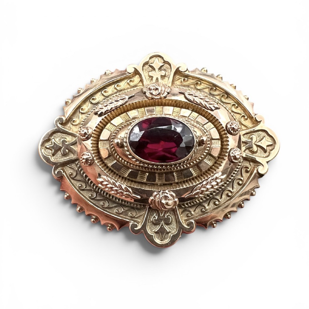 An ornate oval Victorian Etruscan Revival gold brooch featuring a central oval-cut deep red garnet surrounded by detailed relief metalwork and beaded borders. View II