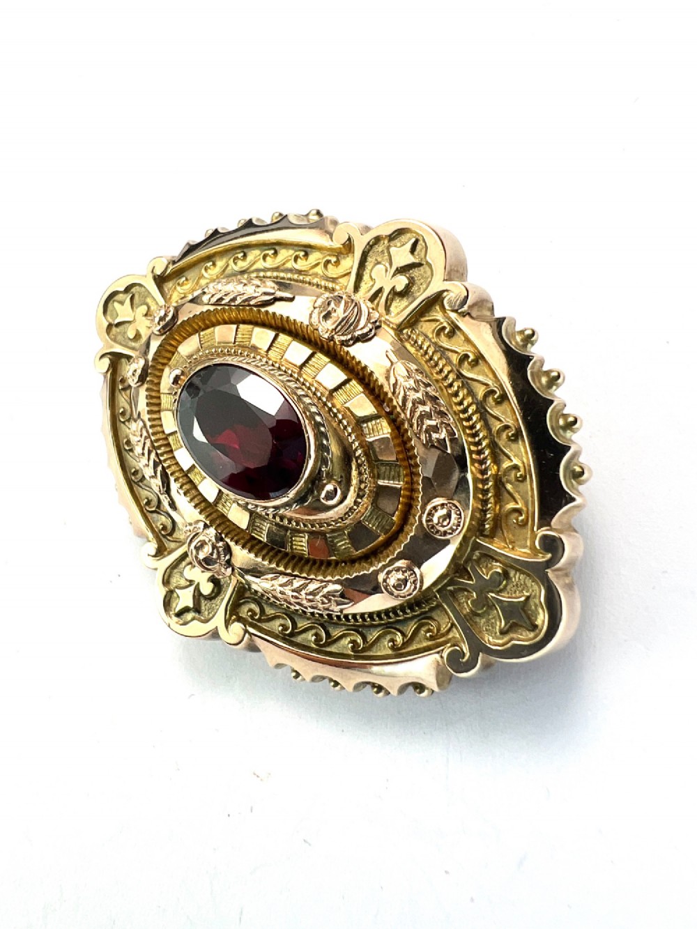 An ornate oval Victorian Etruscan Revival gold brooch featuring a central oval-cut deep red garnet surrounded by detailed relief metalwork and beaded borders. View III