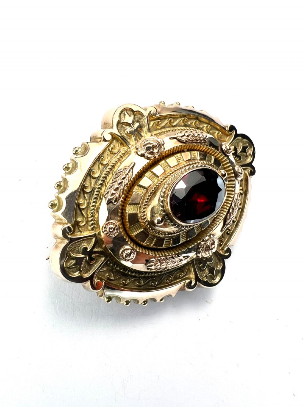 An ornate oval Victorian Etruscan Revival gold brooch featuring a central oval-cut deep red garnet surrounded by detailed relief metalwork and beaded borders. View IV