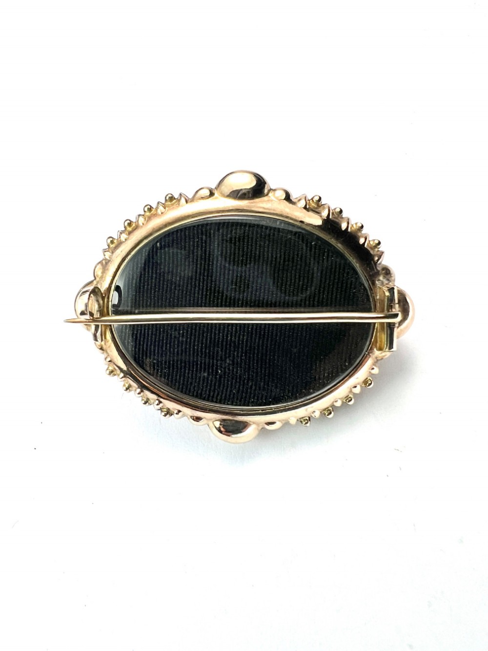 An ornate oval Victorian Etruscan Revival gold brooch featuring a central oval-cut deep red garnet surrounded by detailed relief metalwork and beaded borders. View V