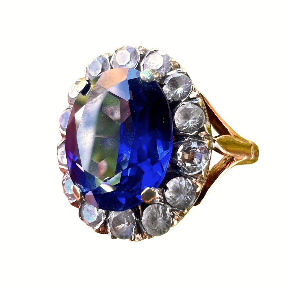 An Edwardian sapphire halo ring featuring a large oval blue sapphire encircled by a halo of clear accent stones set in silver with a yellow gold shank.