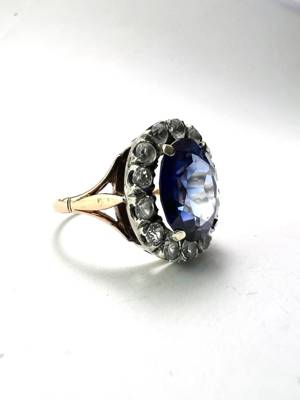 An Edwardian sapphire halo ring featuring a large oval blue sapphire encircled by a halo of clear accent stones set in silver with a yellow gold shank. View V