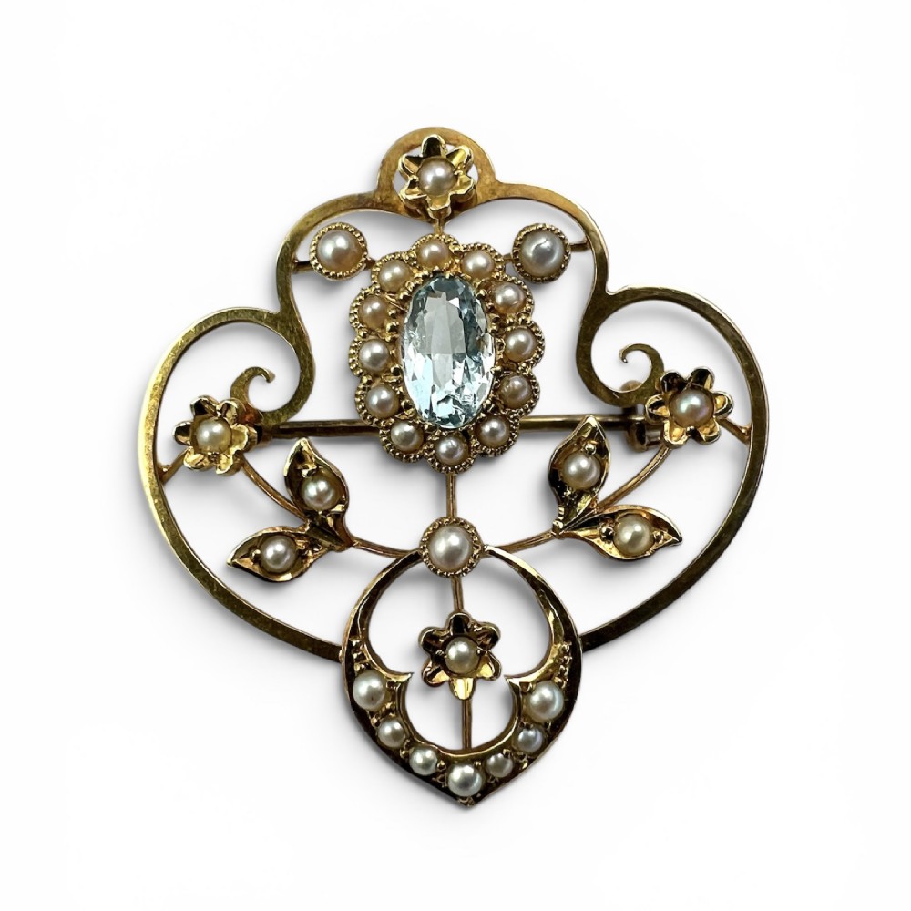 An Edwardian openwork 15ct yellow gold brooch featuring a central oval light blue aquamarine surrounded by seed pearls and floral scroll motifs.