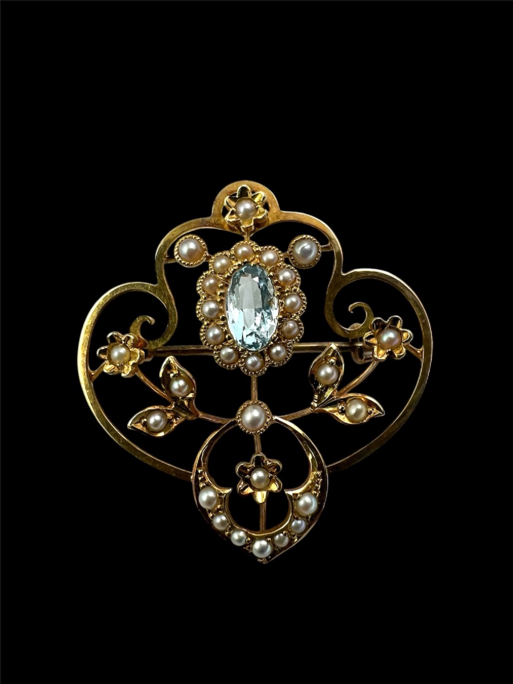 An Edwardian openwork 15ct yellow gold brooch featuring a central oval light blue aquamarine surrounded by seed pearls and floral scroll motifs. View II