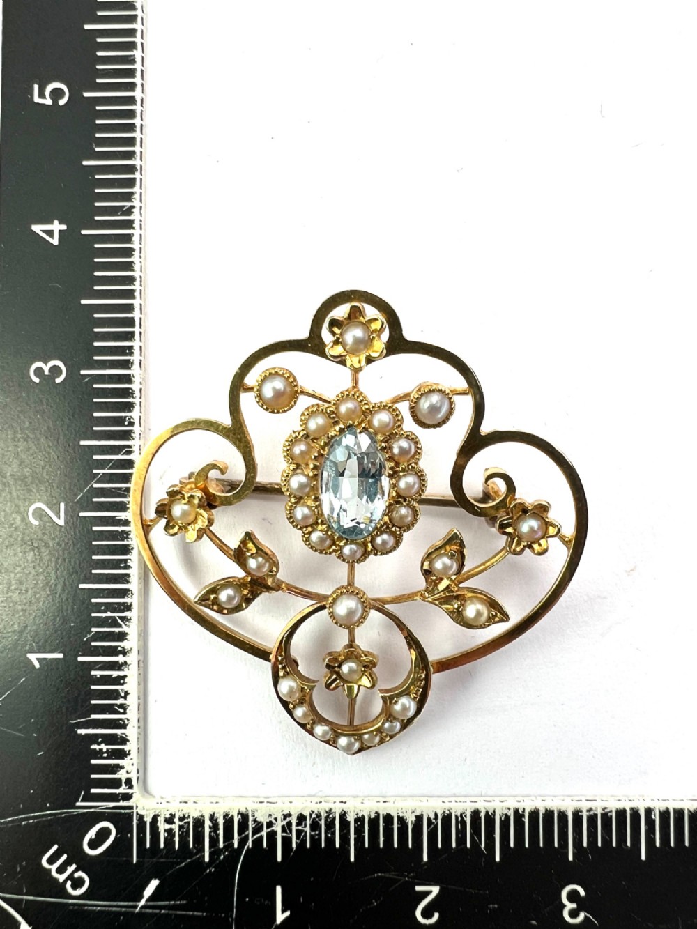 An Edwardian openwork 15ct yellow gold brooch featuring a central oval light blue aquamarine surrounded by seed pearls and floral scroll motifs. View VI