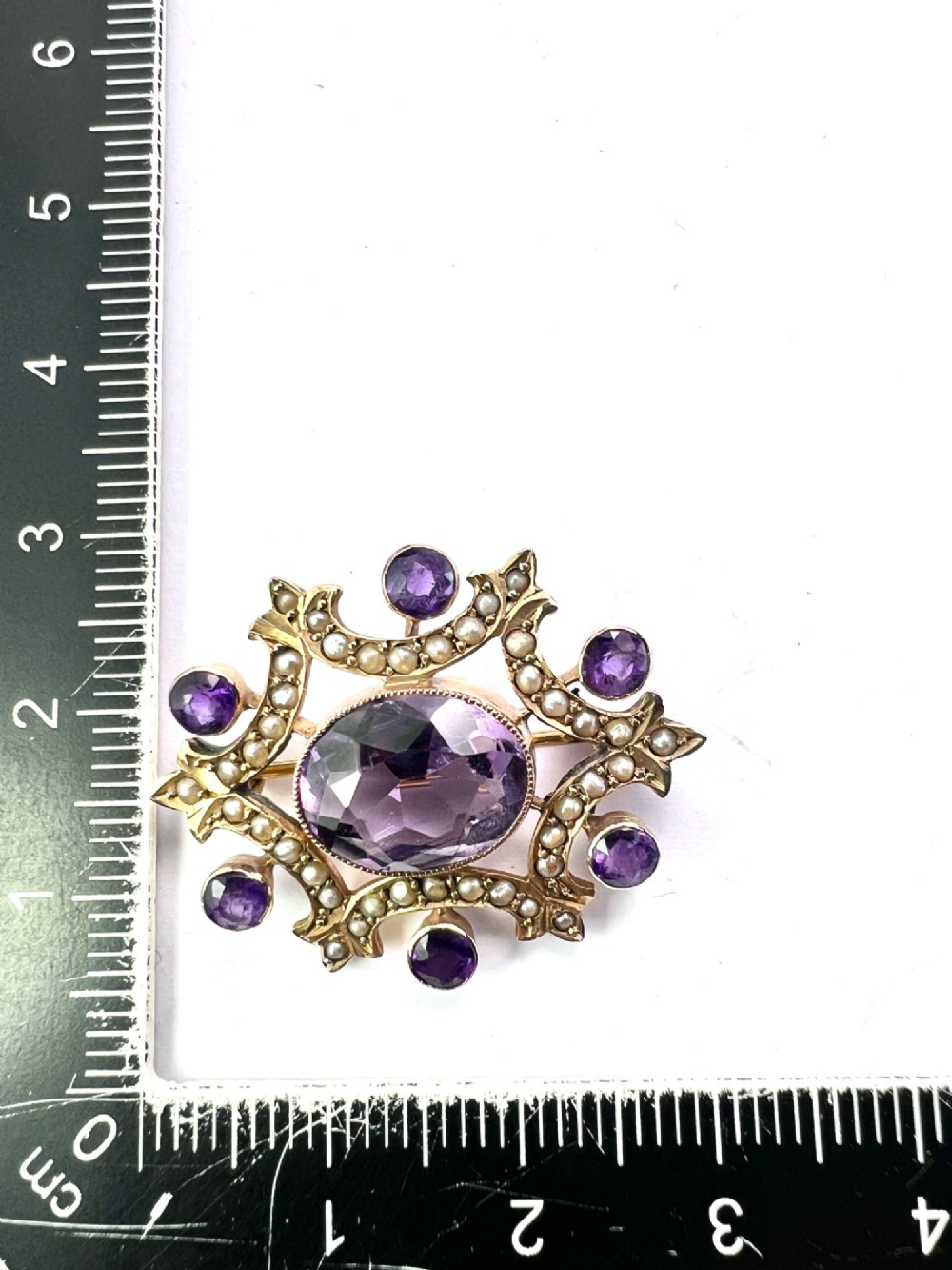 An Edwardian openwork 9ct gold brooch featuring a central oval faceted amethyst surrounded by split seed pearls and six outer round amethysts. View V