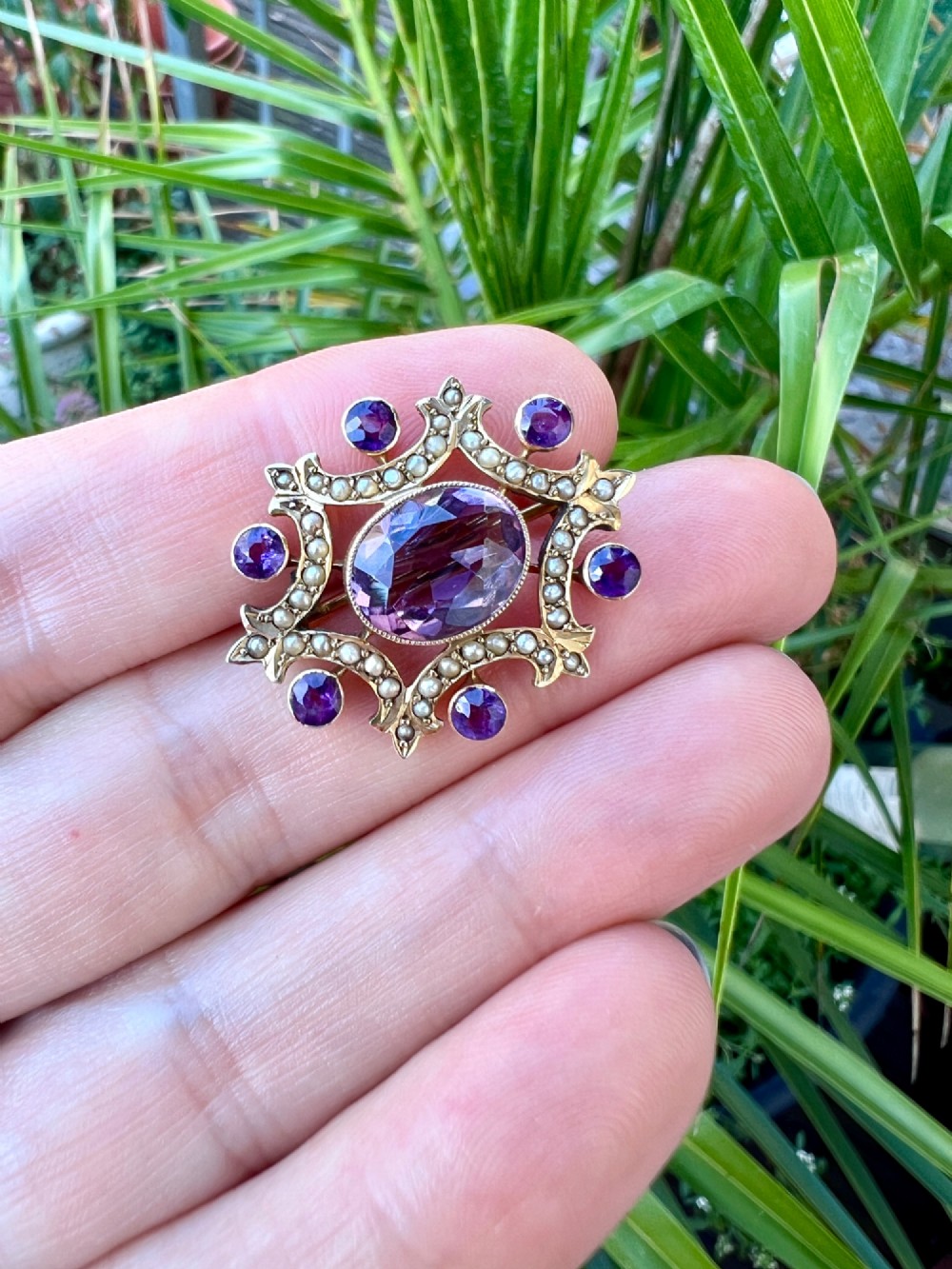 An Edwardian openwork 9ct gold brooch featuring a central oval faceted amethyst surrounded by split seed pearls and six outer round amethysts. View VI