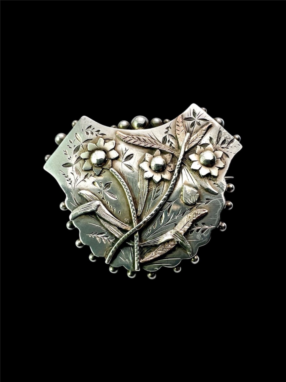 A Victorian silver shield-shaped brooch decorated with applied rose gold flowers, hand engraving, and a beaded outer rim.