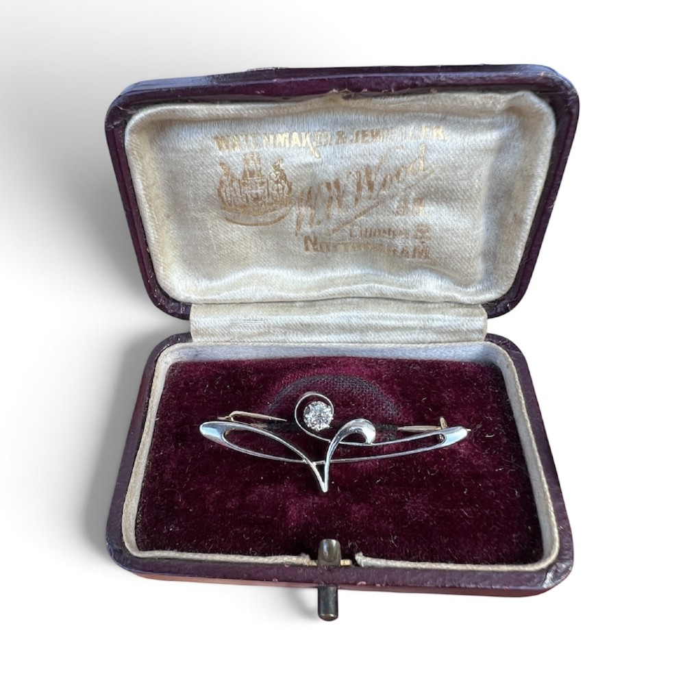 An Art Nouveau 15ct gold and platinum brooch featuring a central diamond set within fluid curvilinear metalwork, presented inside a fitted burgundy velvet box.