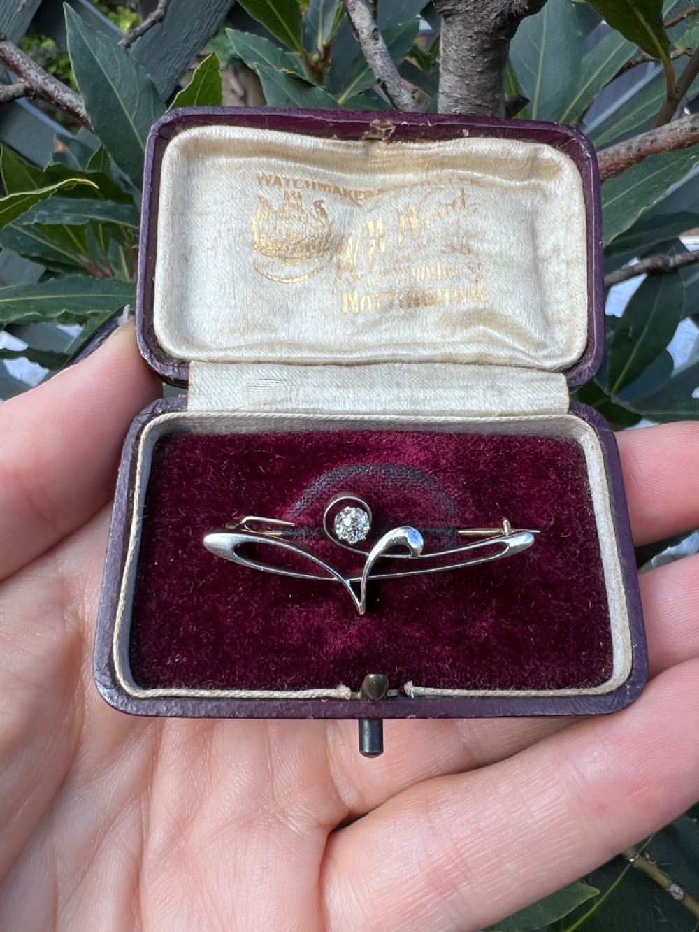 An Art Nouveau 15ct gold and platinum brooch featuring a central diamond set within fluid curvilinear metalwork, presented inside a fitted burgundy velvet box. View VI