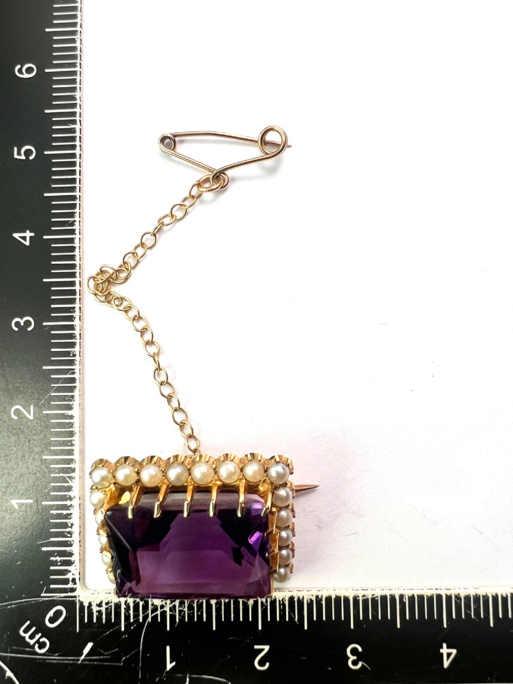 A rectangular Victorian brooch featuring a central facet-cut purple amethyst encircled by a halo of small white pearls set in 15ct gold with an attached safety chain. View V