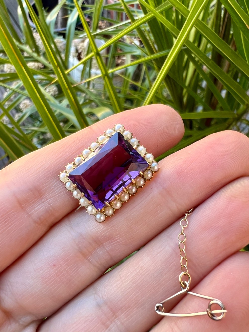 A rectangular Victorian brooch featuring a central facet-cut purple amethyst encircled by a halo of small white pearls set in 15ct gold with an attached safety chain. View VI