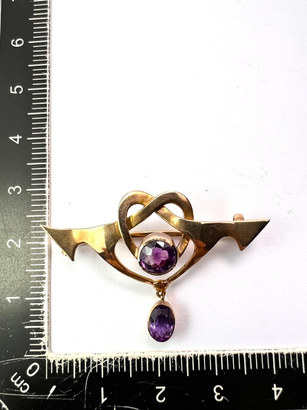 An Art Nouveau 9ct gold brooch featuring a heart-shaped whiplash frame set with a round purple amethyst and a suspended amethyst drop, displayed inside a fitted velvet case. View V