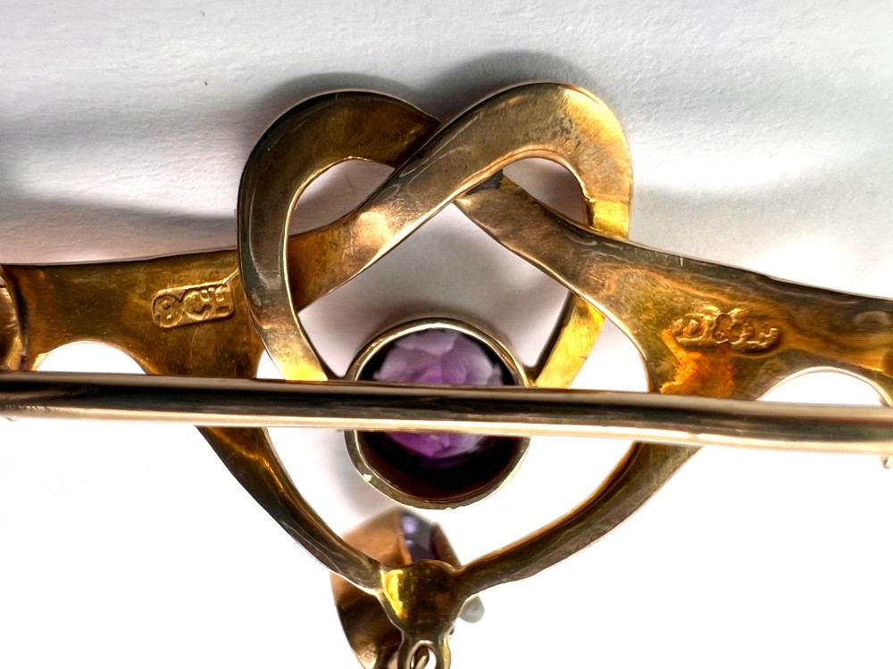 An Art Nouveau 9ct gold brooch featuring a heart-shaped whiplash frame set with a round purple amethyst and a suspended amethyst drop, displayed inside a fitted velvet case. View VI