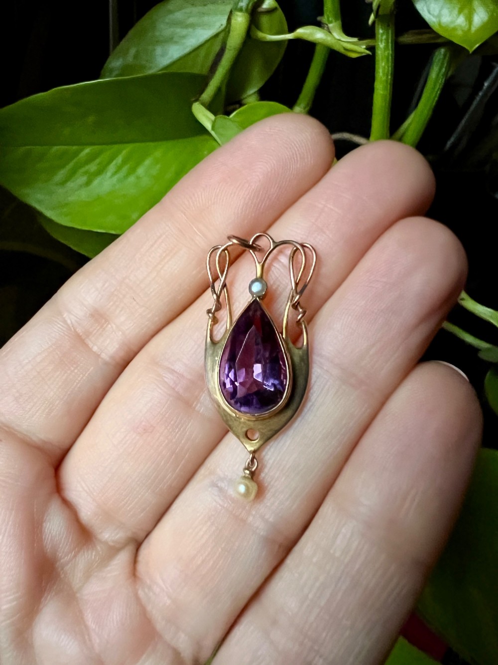 An Art Nouveau gold pendant features an openwork whiplash frame surrounding a central pear-cut purple amethyst gemstone, anchored by a small pearl above and a dangling seed pearl below. View IV