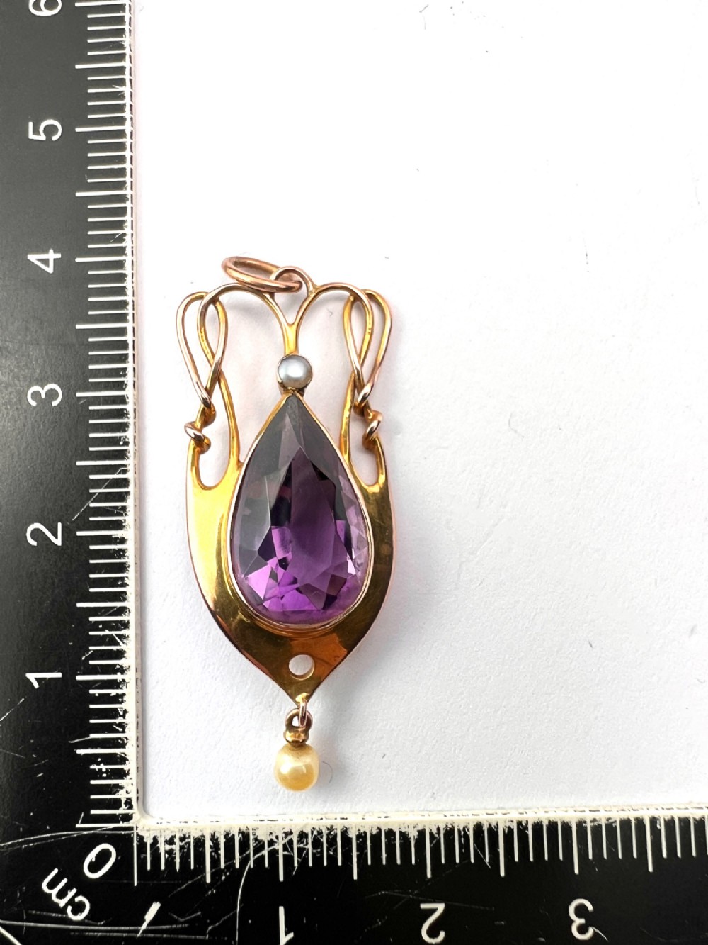 An Art Nouveau gold pendant features an openwork whiplash frame surrounding a central pear-cut purple amethyst gemstone, anchored by a small pearl above and a dangling seed pearl below. View V