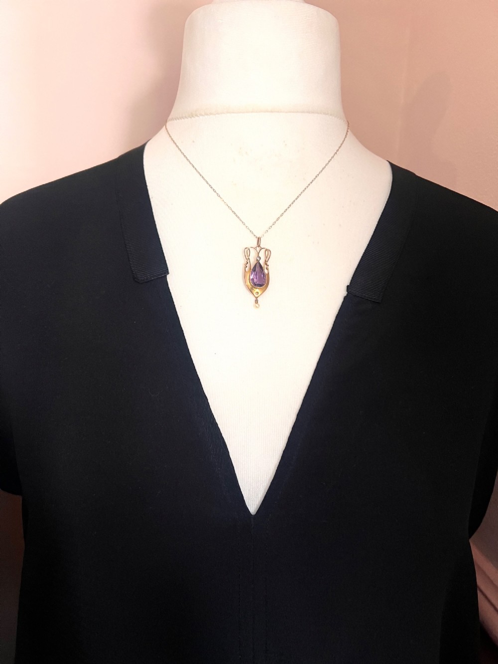 An Art Nouveau gold pendant features an openwork whiplash frame surrounding a central pear-cut purple amethyst gemstone, anchored by a small pearl above and a dangling seed pearl below. View VI
