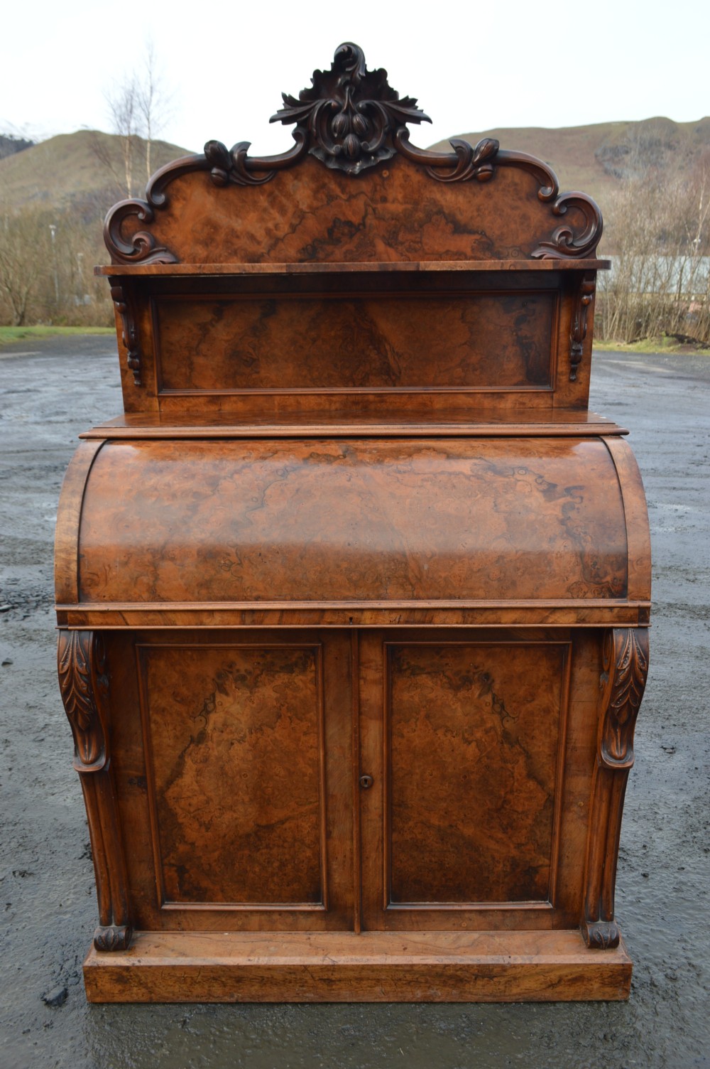 A Victorian burr walnut cylinder front bureau with a carved top pediment, a raised shelf, a curved roll front, and lower cupboard doors.