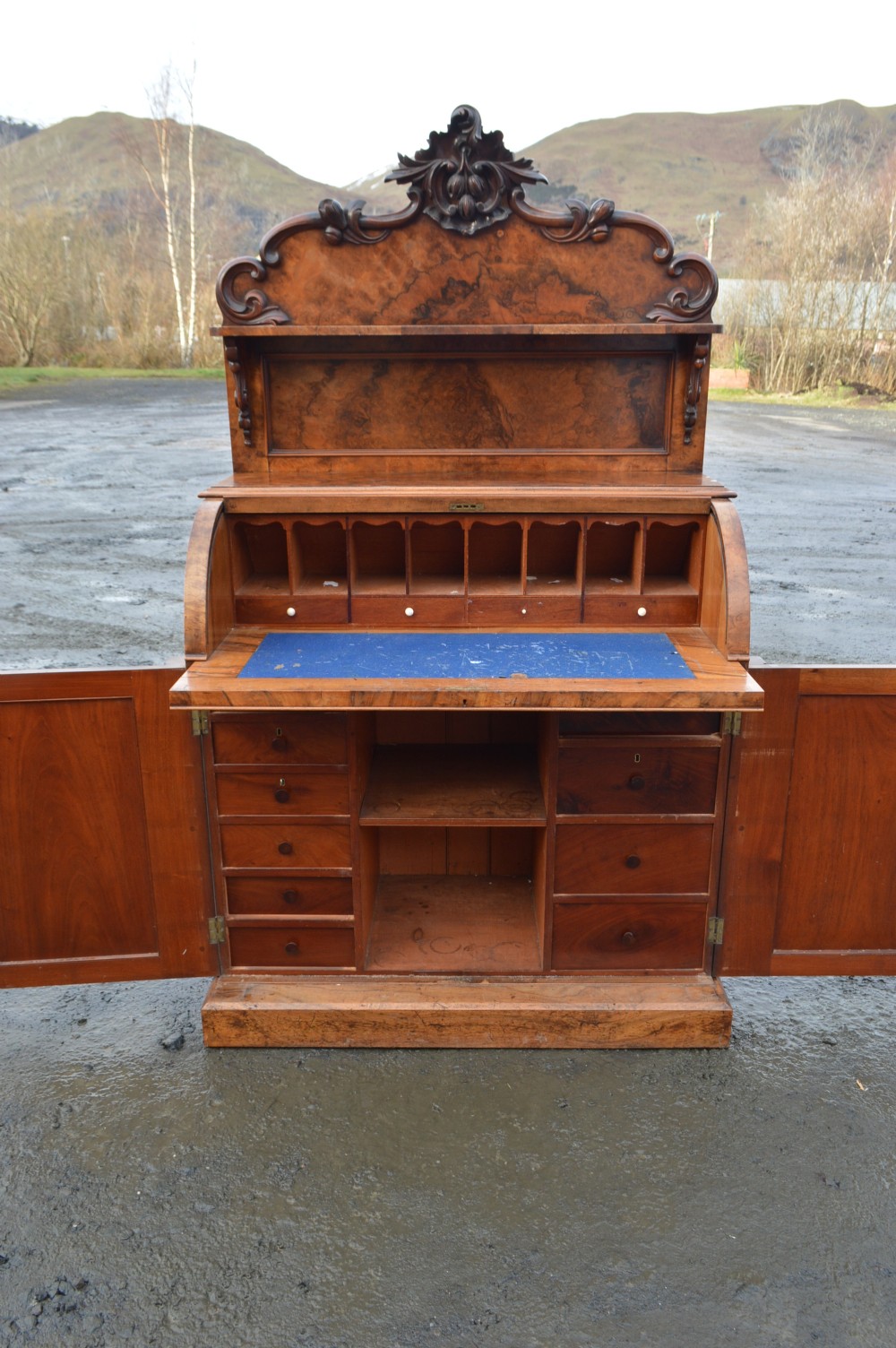 A Victorian burr walnut cylinder front bureau with a carved top pediment, a raised shelf, a curved roll front, and lower cupboard doors. View IV