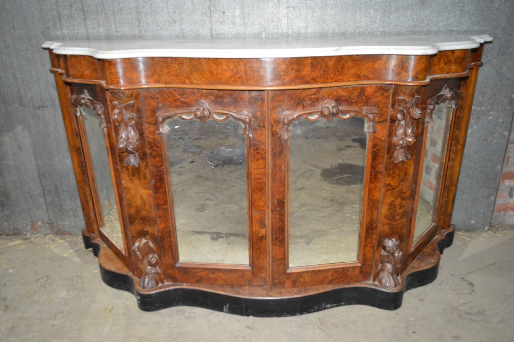 A Victorian serpentine burr walnut credenza featuring a white shaped marble top, four mirrored doors, and carved foliate pilasters.