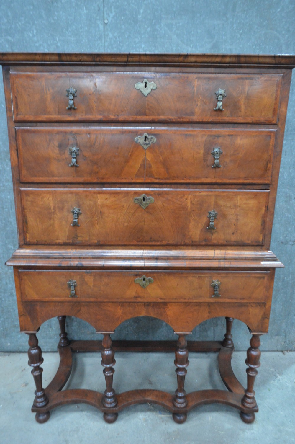A George I walnut chest on stand featuring multiple veneered drawers with brass drop handles, an arched lower apron, and turned legs connected by a wavy stretcher.