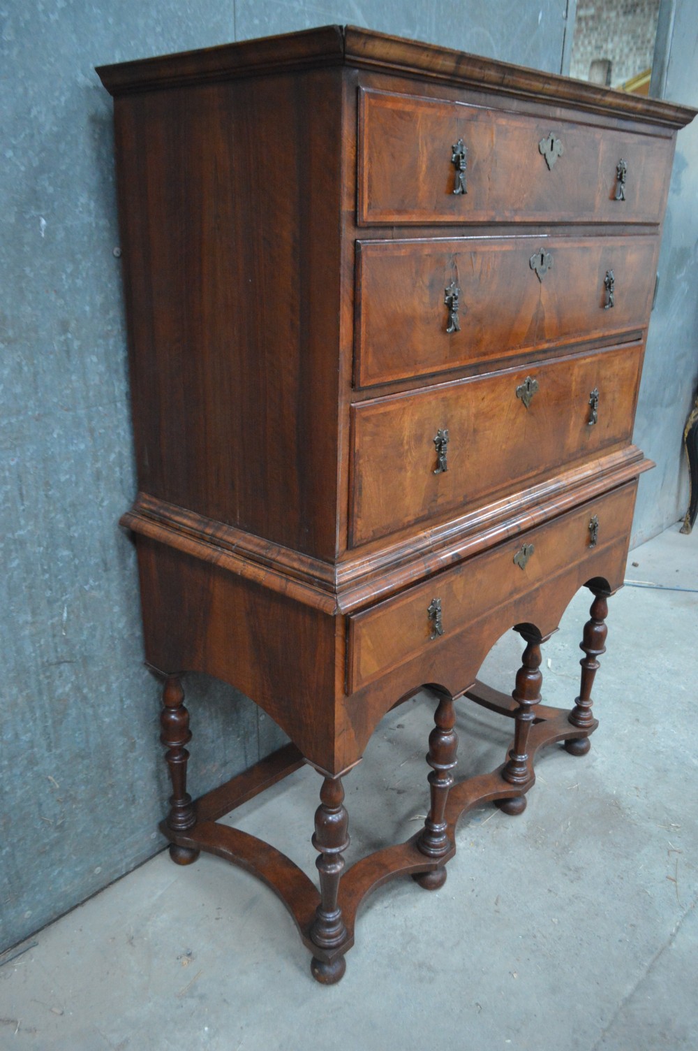 A George I walnut chest on stand featuring multiple veneered drawers with brass drop handles, an arched lower apron, and turned legs connected by a wavy stretcher. View III