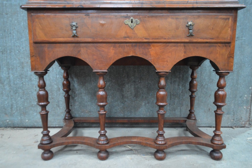 A George I walnut chest on stand featuring multiple veneered drawers with brass drop handles, an arched lower apron, and turned legs connected by a wavy stretcher. View V