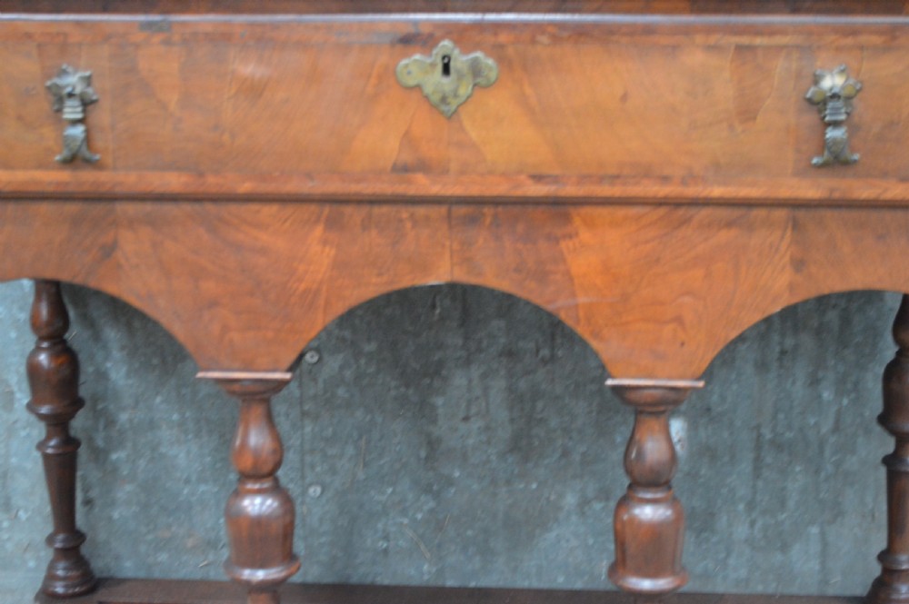 A George I walnut chest on stand featuring multiple veneered drawers with brass drop handles, an arched lower apron, and turned legs connected by a wavy stretcher. View VI