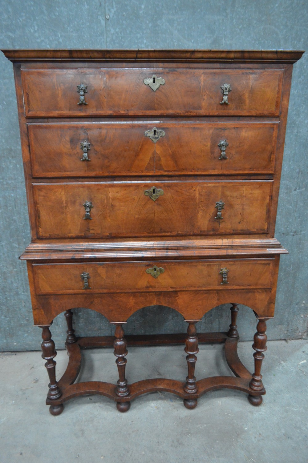A George I walnut chest on stand featuring multiple veneered drawers with brass drop handles, an arched lower apron, and turned legs connected by a wavy stretcher. View VII