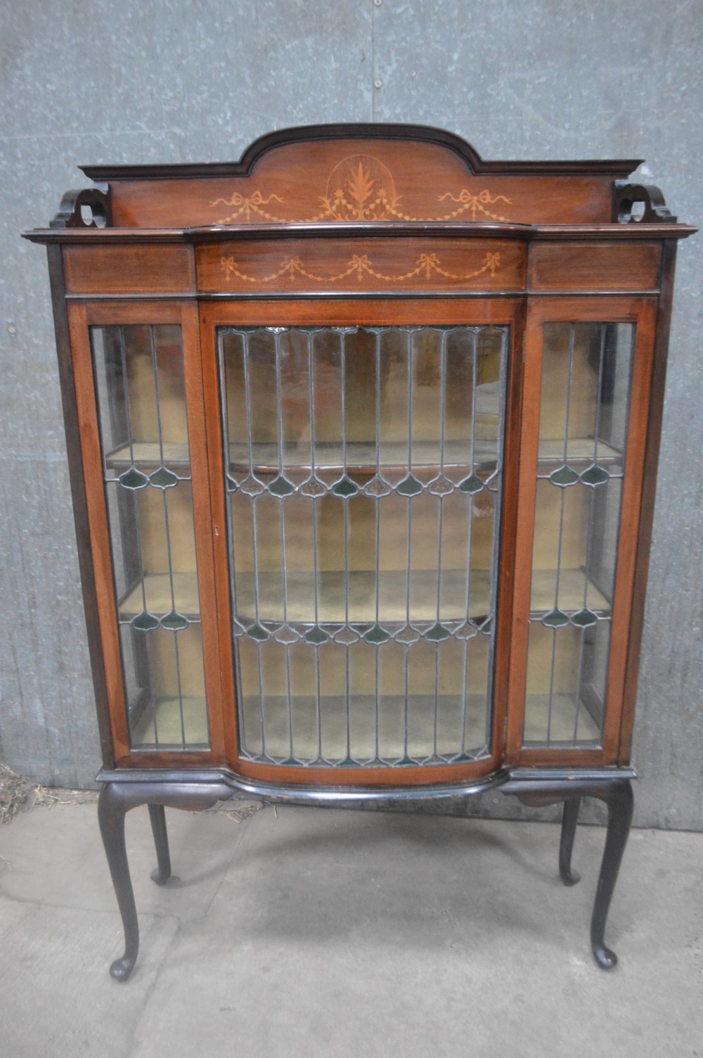 An Edwardian mahogany display cabinet featuring inlaid satinwood swags on the pediment, leaded glass doors with green accents, and slender cabriole legs.
