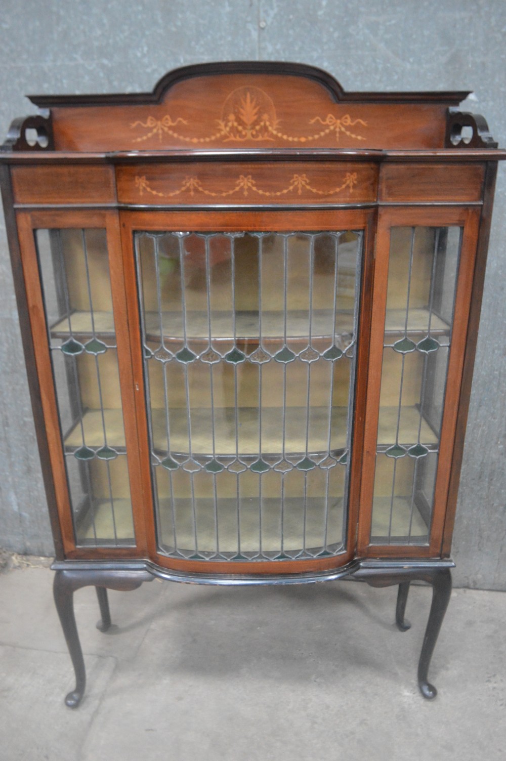 An Edwardian mahogany display cabinet featuring inlaid satinwood swags on the pediment, leaded glass doors with green accents, and slender cabriole legs. View VI