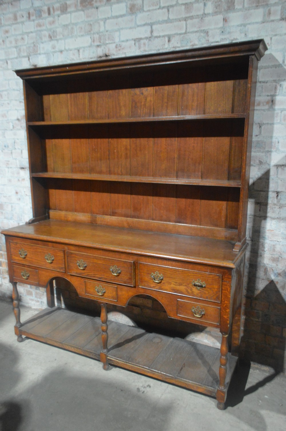 A tall Victorian oak plate dresser featuring a three-tier open plate rack mounted above a five-drawer base with brass swan-neck handles and a potboard shelf below.