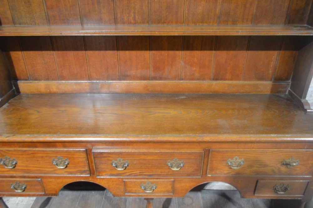 A tall Victorian oak plate dresser featuring a three-tier open plate rack mounted above a five-drawer base with brass swan-neck handles and a potboard shelf below. View II