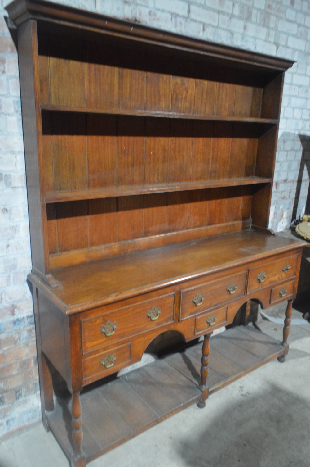 A tall Victorian oak plate dresser featuring a three-tier open plate rack mounted above a five-drawer base with brass swan-neck handles and a potboard shelf below. View III