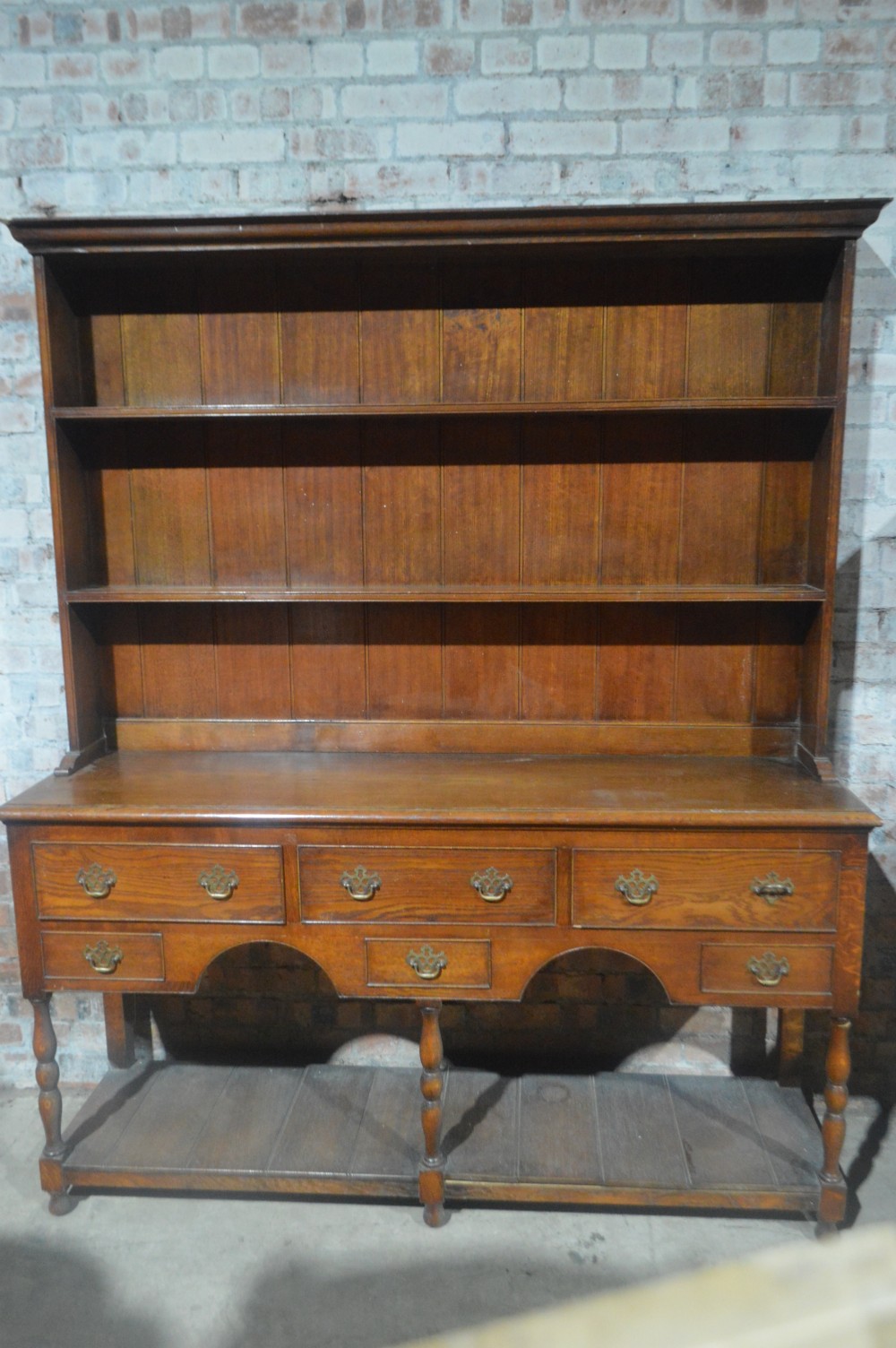 A tall Victorian oak plate dresser featuring a three-tier open plate rack mounted above a five-drawer base with brass swan-neck handles and a potboard shelf below. View IV