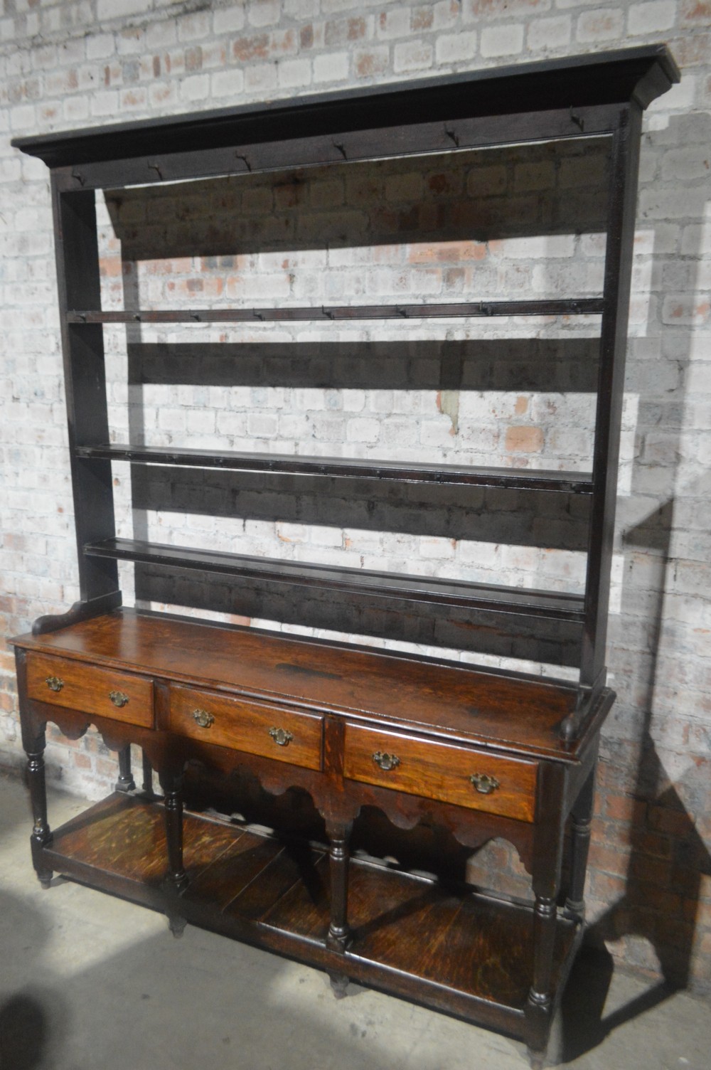 A tall dark oak Victorian dresser featuring an upper open plate rack with hooks above three brass-handled drawers, a scalloped apron, and a lower potboard shelf.