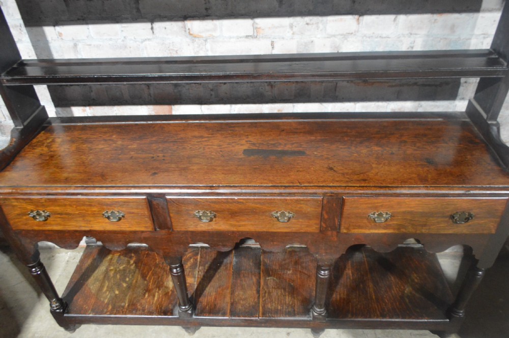 A tall dark oak Victorian dresser featuring an upper open plate rack with hooks above three brass-handled drawers, a scalloped apron, and a lower potboard shelf. View II