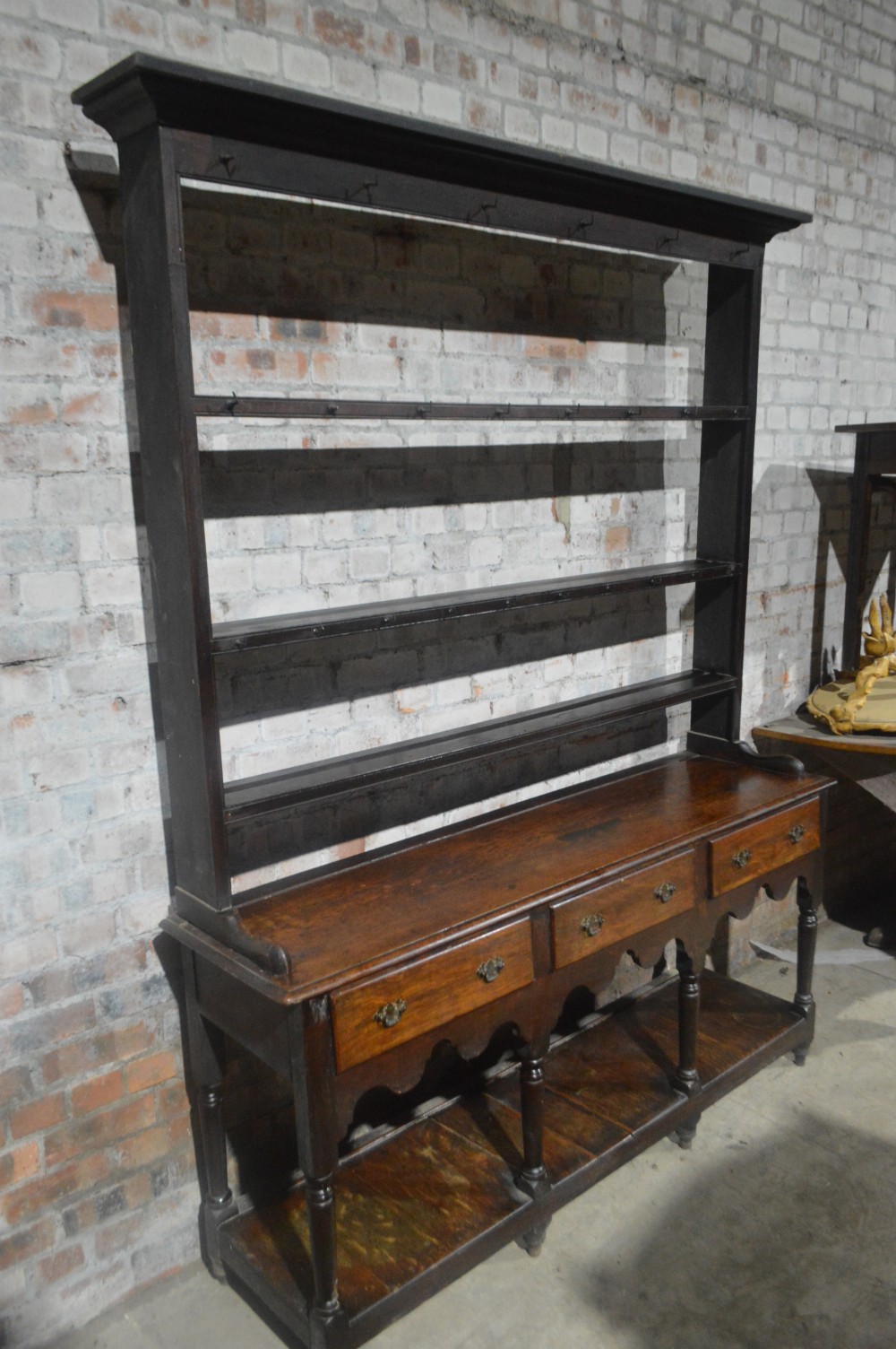 A tall dark oak Victorian dresser featuring an upper open plate rack with hooks above three brass-handled drawers, a scalloped apron, and a lower potboard shelf. View V