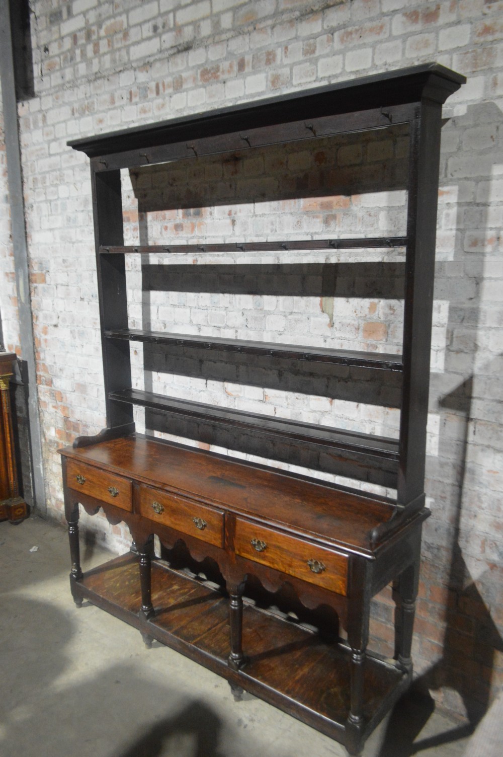 A tall dark oak Victorian dresser featuring an upper open plate rack with hooks above three brass-handled drawers, a scalloped apron, and a lower potboard shelf. View VI