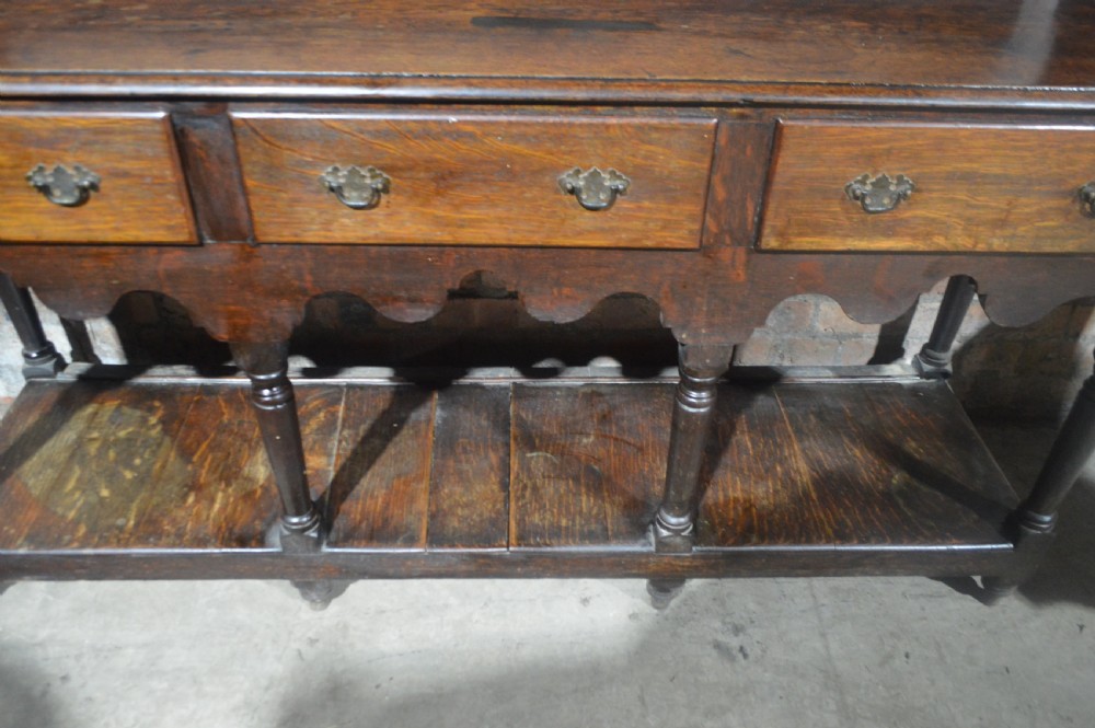 A tall dark oak Victorian dresser featuring an upper open plate rack with hooks above three brass-handled drawers, a scalloped apron, and a lower potboard shelf. View VII
