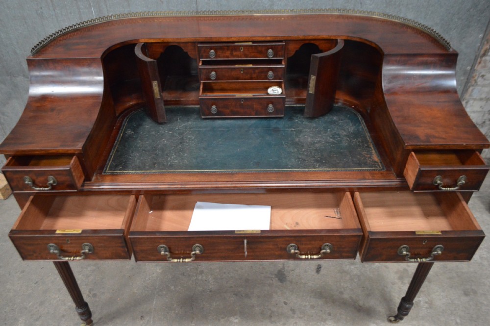 An Edwardian mahogany Carlton House writing desk featuring a curved U-shaped superstructure with small drawers, pigeonholes, a brass gallery, a dark leather insert, and fluted legs on brass castors. View VI