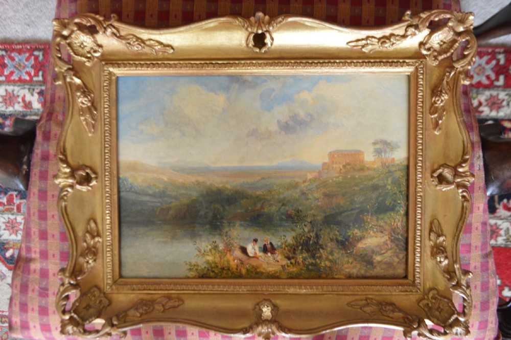 A 19th-century oil painting depicting a countryside river landscape with two seated figures in the foreground and a large building on a hill in the distance under a cloudy sky. View II