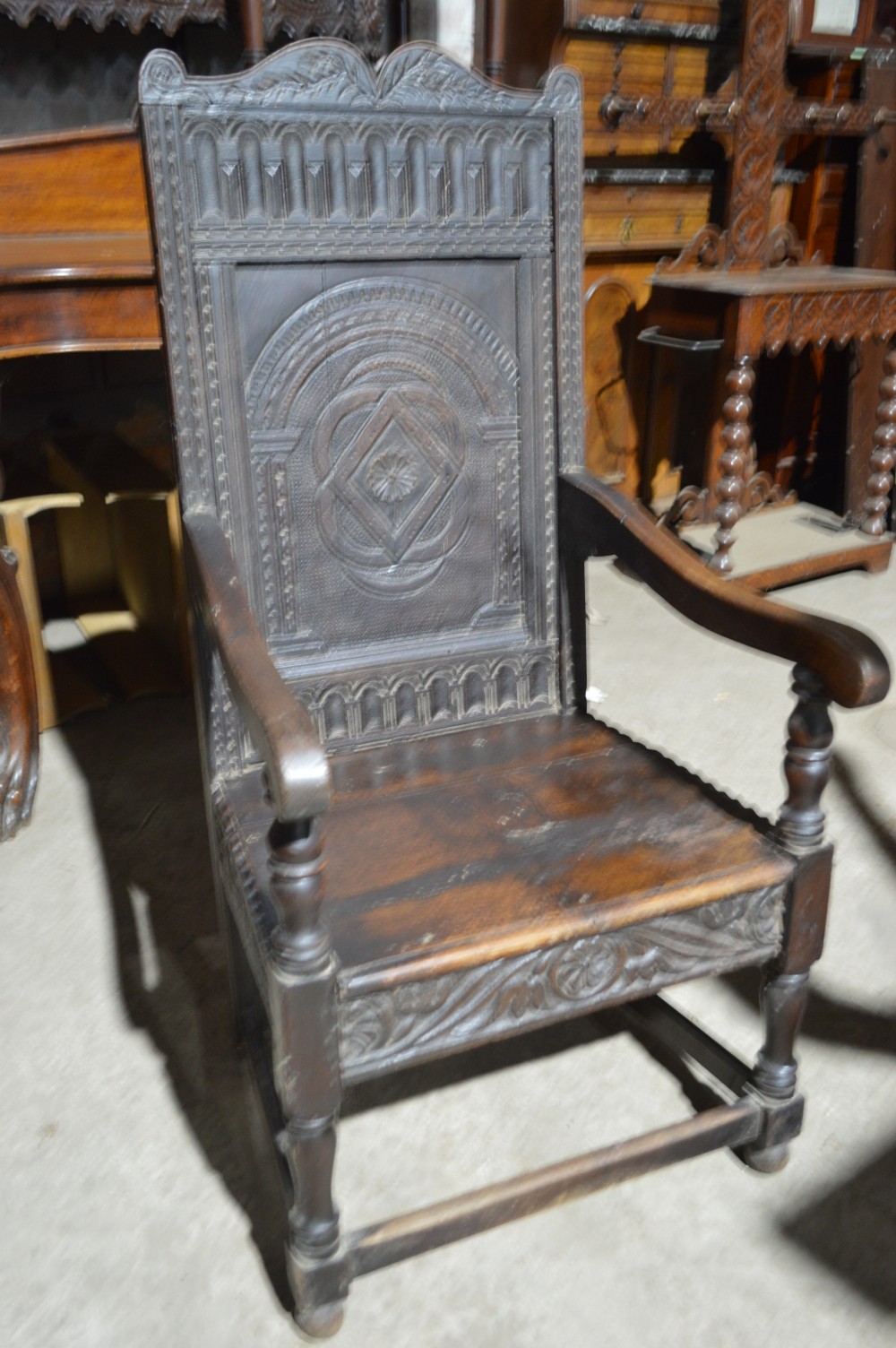 A dark carved oak wainscot armchair featuring a detailed arched panel back, turned front legs, and sturdy stretchers.