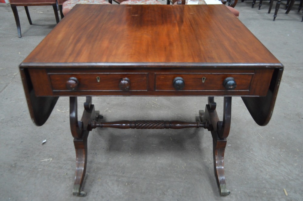 A Georgian mahogany sofa table featuring twin drop leaves, two front drawers with round wooden knobs, a turned cross-stretcher, and splayed legs ending in brass caps.