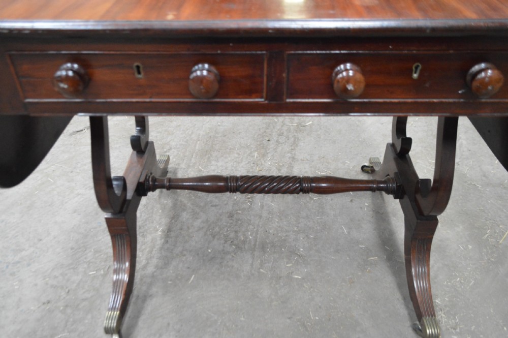 A Georgian mahogany sofa table featuring twin drop leaves, two front drawers with round wooden knobs, a turned cross-stretcher, and splayed legs ending in brass caps. View VI