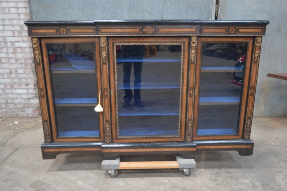 A Victorian ebonised and amboyna three-door breakfront glazed bookcase featuring intricate inlays, brass mounts, and a blue-lined interior.