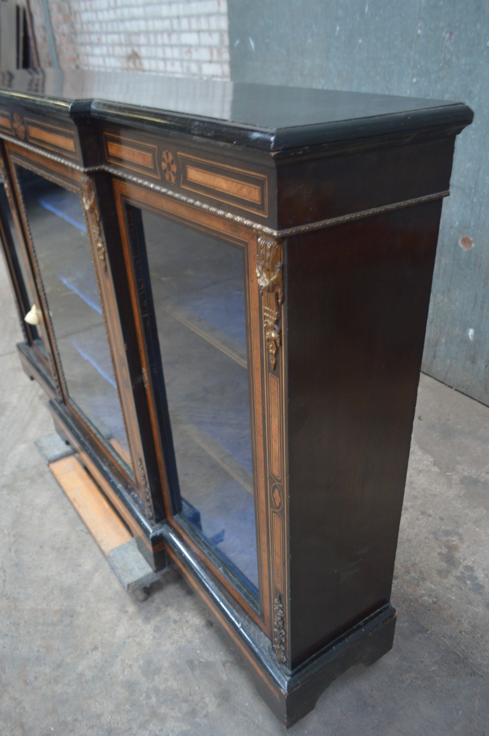 A Victorian ebonised and amboyna three-door breakfront glazed bookcase featuring intricate inlays, brass mounts, and a blue-lined interior. View VI