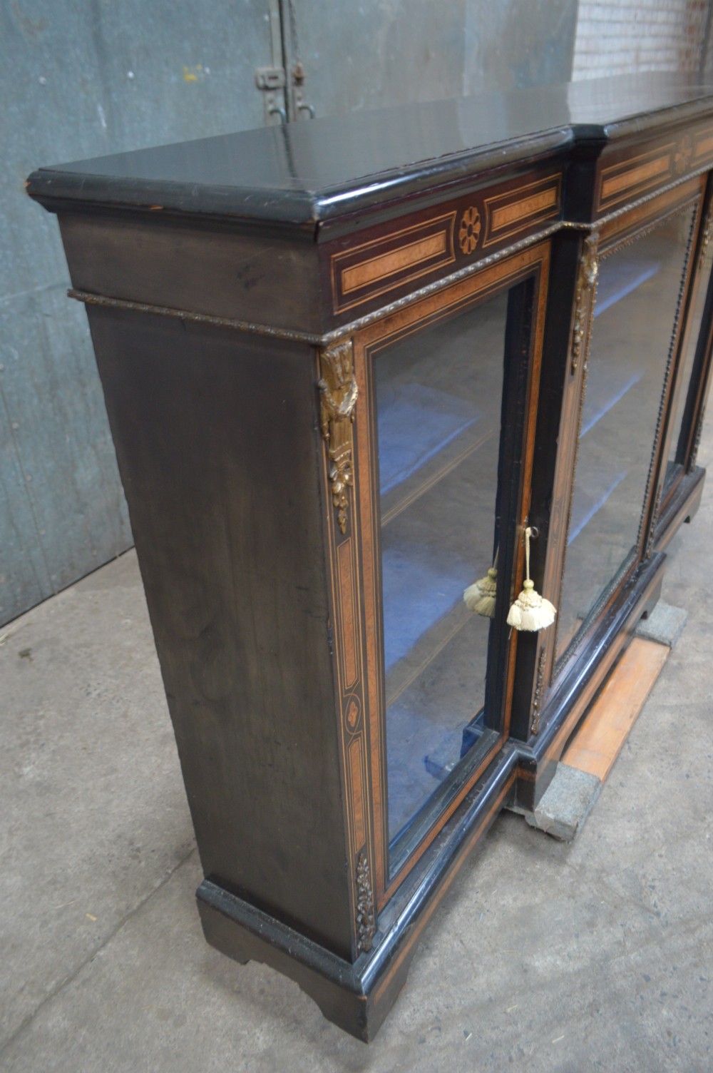 A Victorian ebonised and amboyna three-door breakfront glazed bookcase featuring intricate inlays, brass mounts, and a blue-lined interior. View VII
