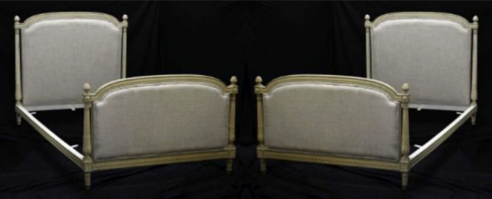 A pair of French Louis XVI style twin bedsteads featuring carved off-white painted wood frames, fluted corner posts with finials, and grey fabric upholstered headboards and footboards.