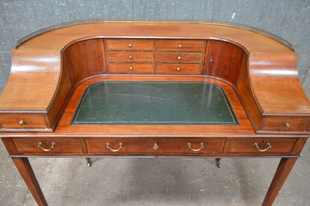 An Edwardian mahogany Carlton House desk featuring a raised U-shaped gallery of drawers, a brass rail, a green leather writing surface, and tapered square legs with castors. View II